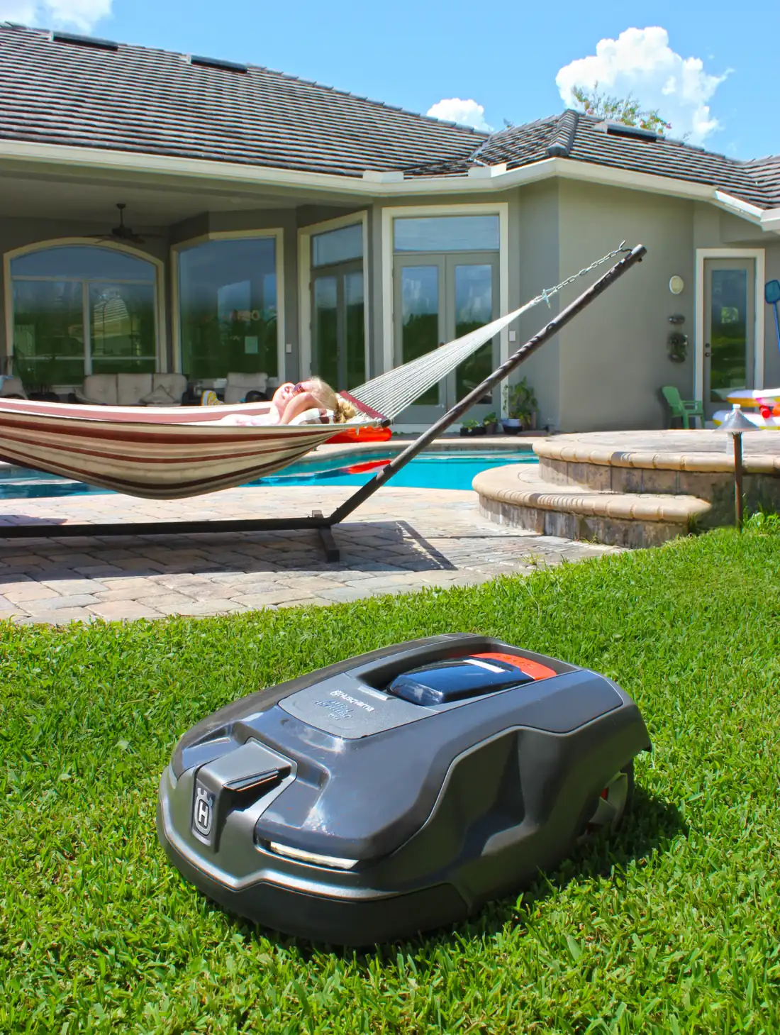 A robotic mower working on a lawn beside a pool while the homeowner relaxes in a hammock